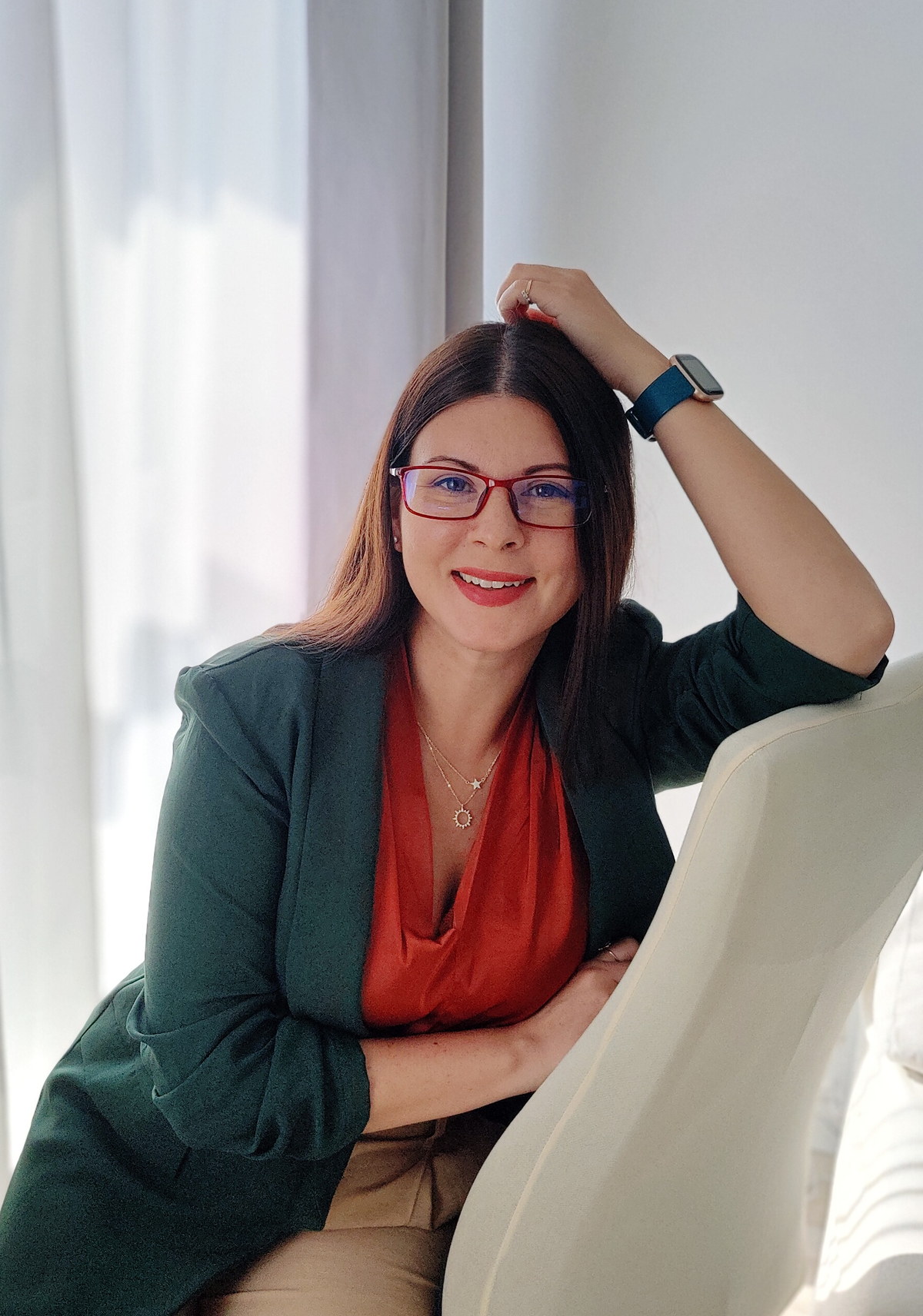 Yuli Varzar smiling, wearing a green blazer and red glasses, seated on a chair